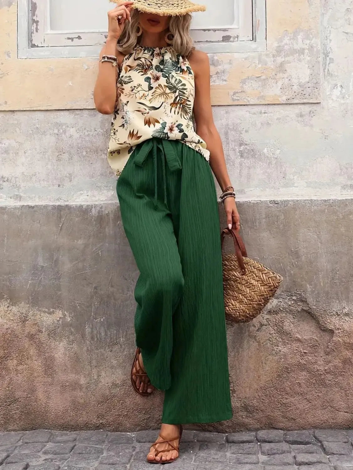 Woman in sleeveless floral top, wide-leg green pants, straw hat, woven bag, and sandals