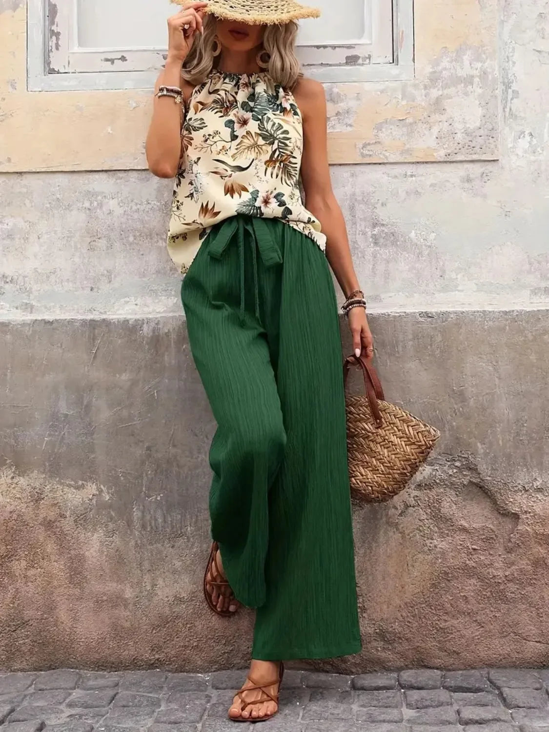 Woman in sleeveless floral top, wide-leg green pants, straw hat, woven bag, and sandals