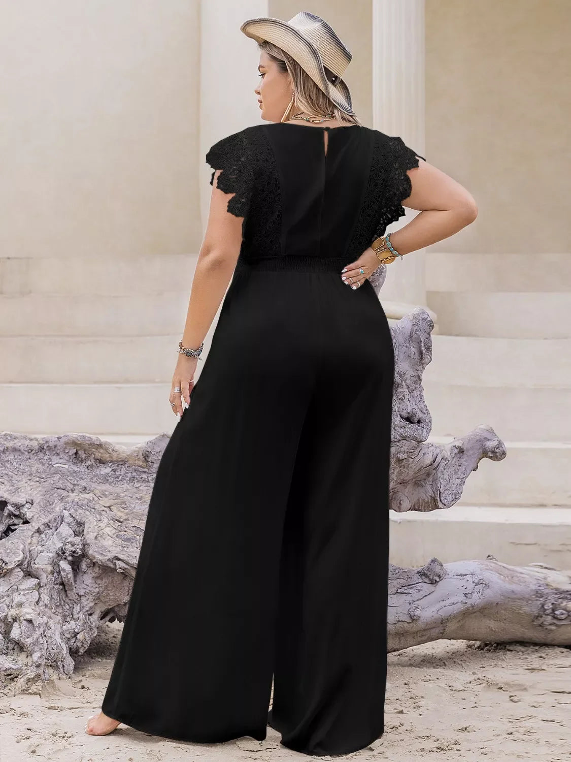 Woman in black wide-leg jumpsuit with lace sleeves and straw hat posing outdoors on sand