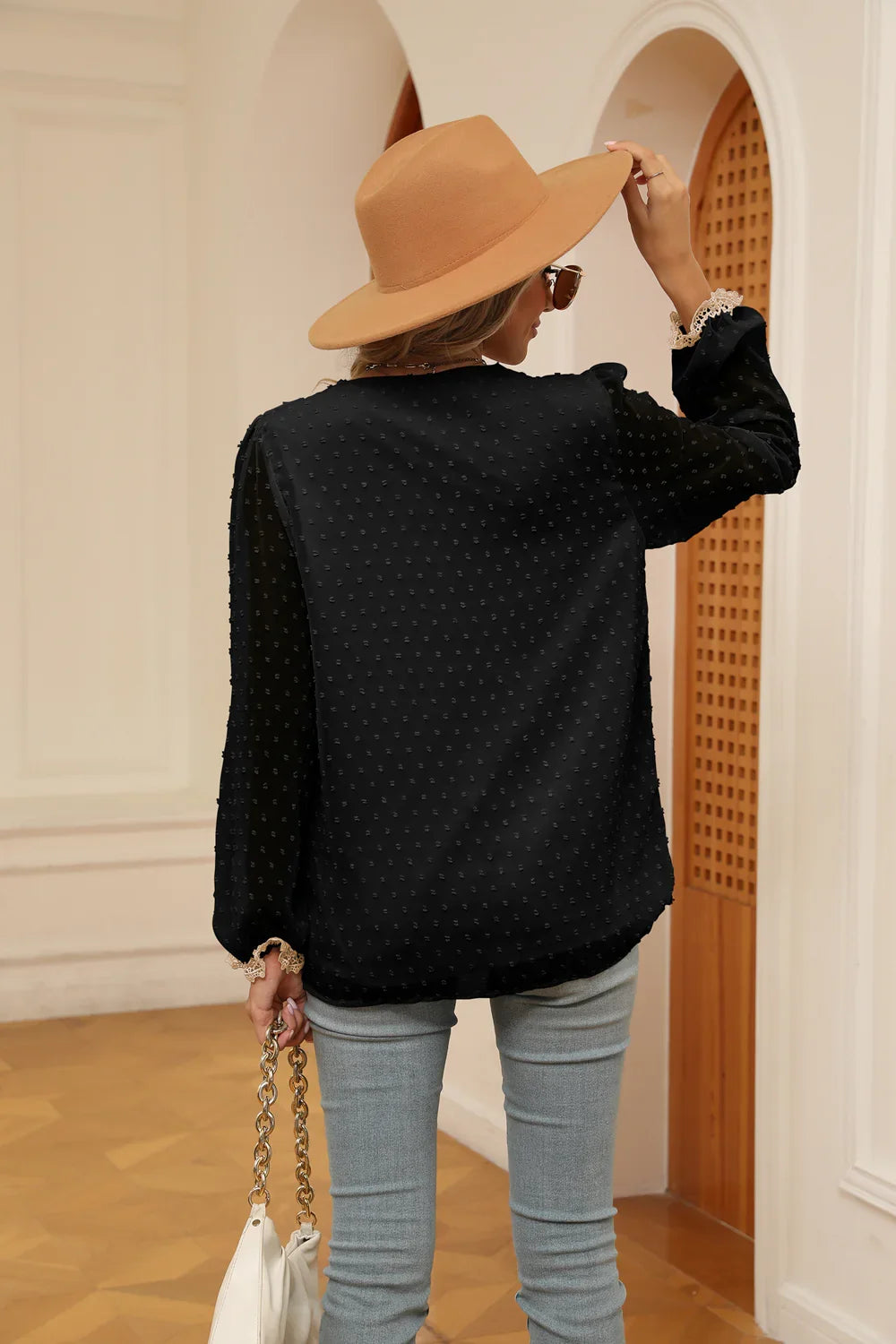 Woman wearing a black textured blouse with lace cuffs, beige wide-brim hat, sunglasses, and light gray jeans indoors