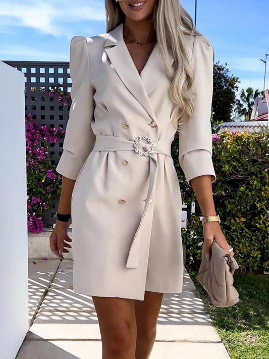 Woman wearing beige belted blazer dress with gold buttons and carrying a matching clutch outdoors
