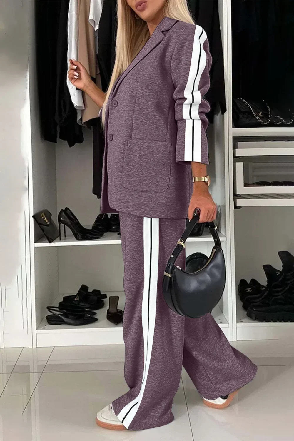 Woman wearing purple track suit with white stripes holding black handbag in stylish closet