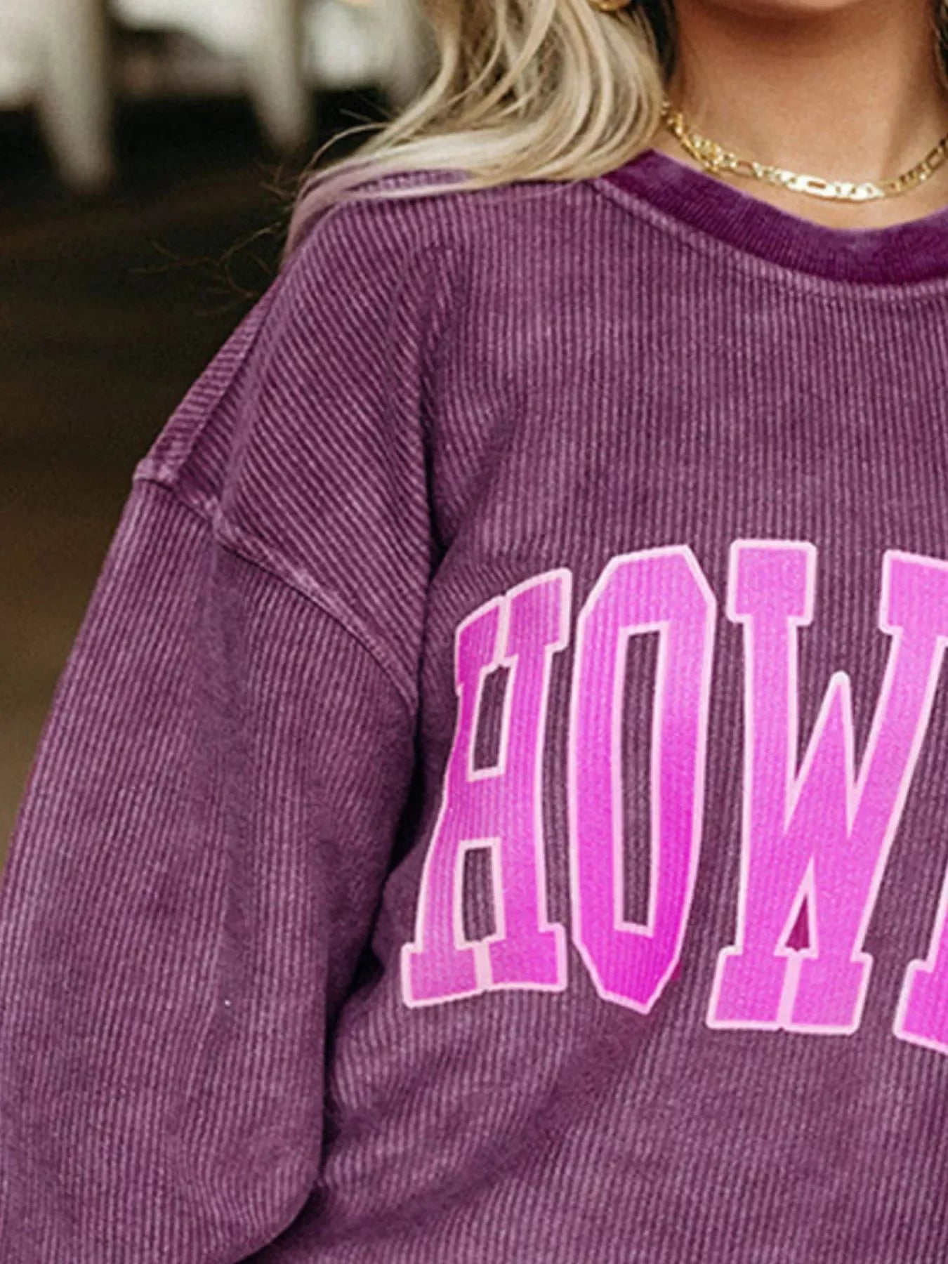 Woman wearing purple ribbed sweatshirt with pink lettering and gold necklace