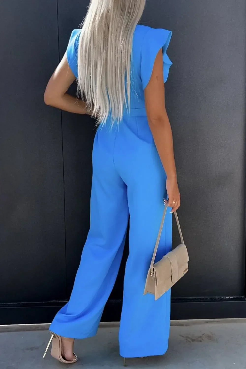 Woman with long blonde hair wearing a blue jumpsuit and beige heels, holding a beige handbag