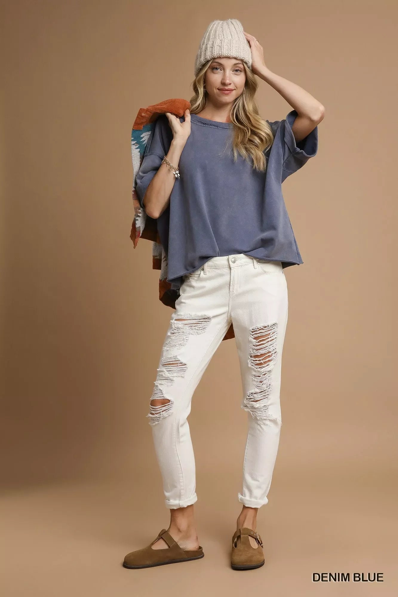Woman in denim blue oversized top, cream ripped jeans, beige knit beanie, and brown slip-on shoes against brown background