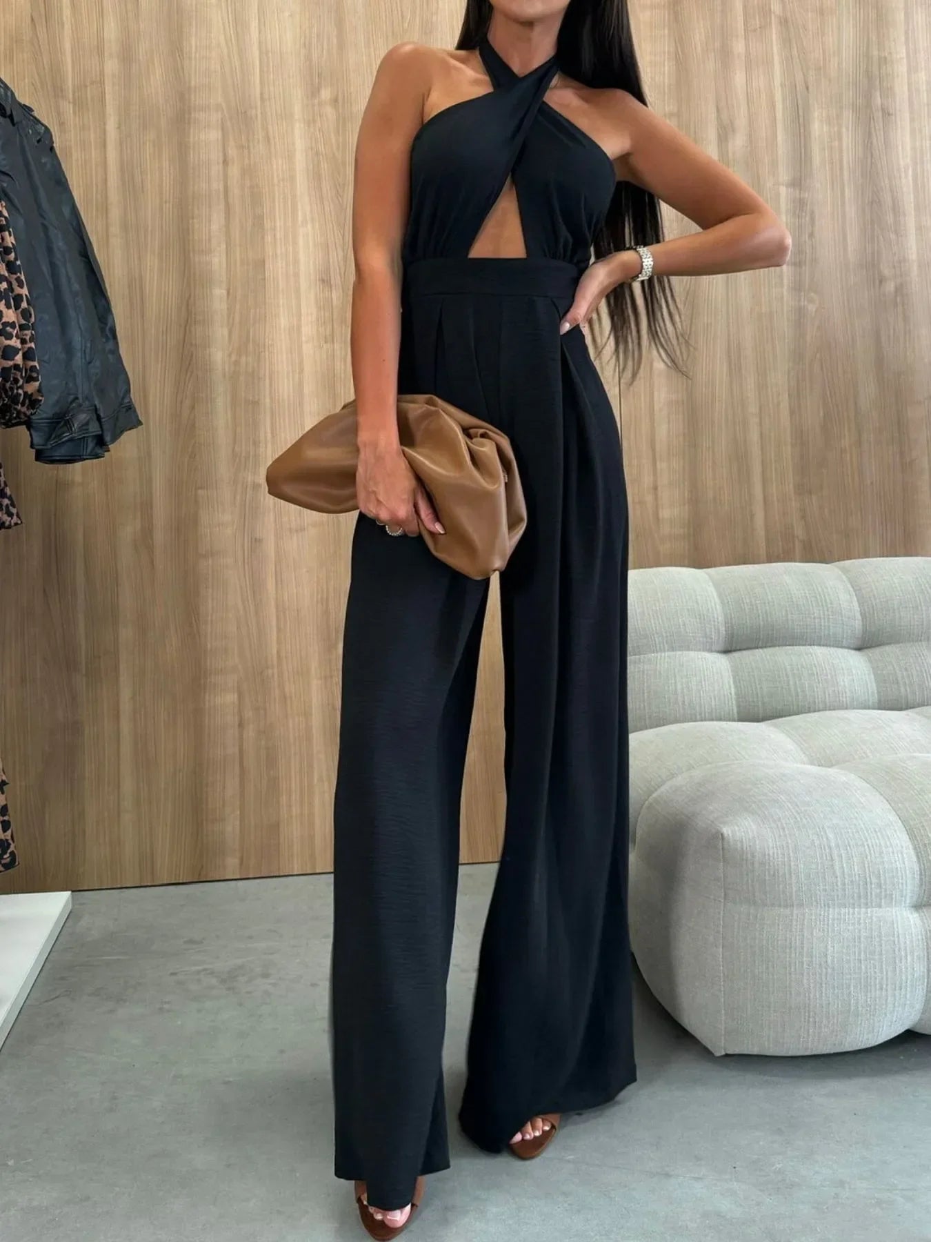 Woman wearing elegant black halter-neck wide-leg jumpsuit holding brown clutch bag indoors