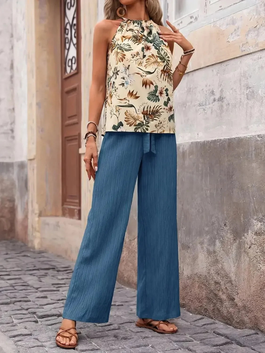 Woman wearing a floral halter top and wide-leg blue pants on a stone street.