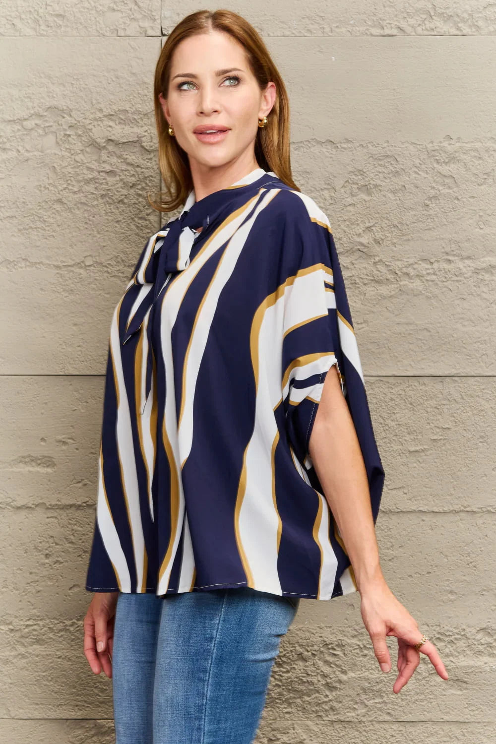 Woman wearing a navy, white, and gold striped blouse with bow tie detail and blue jeans