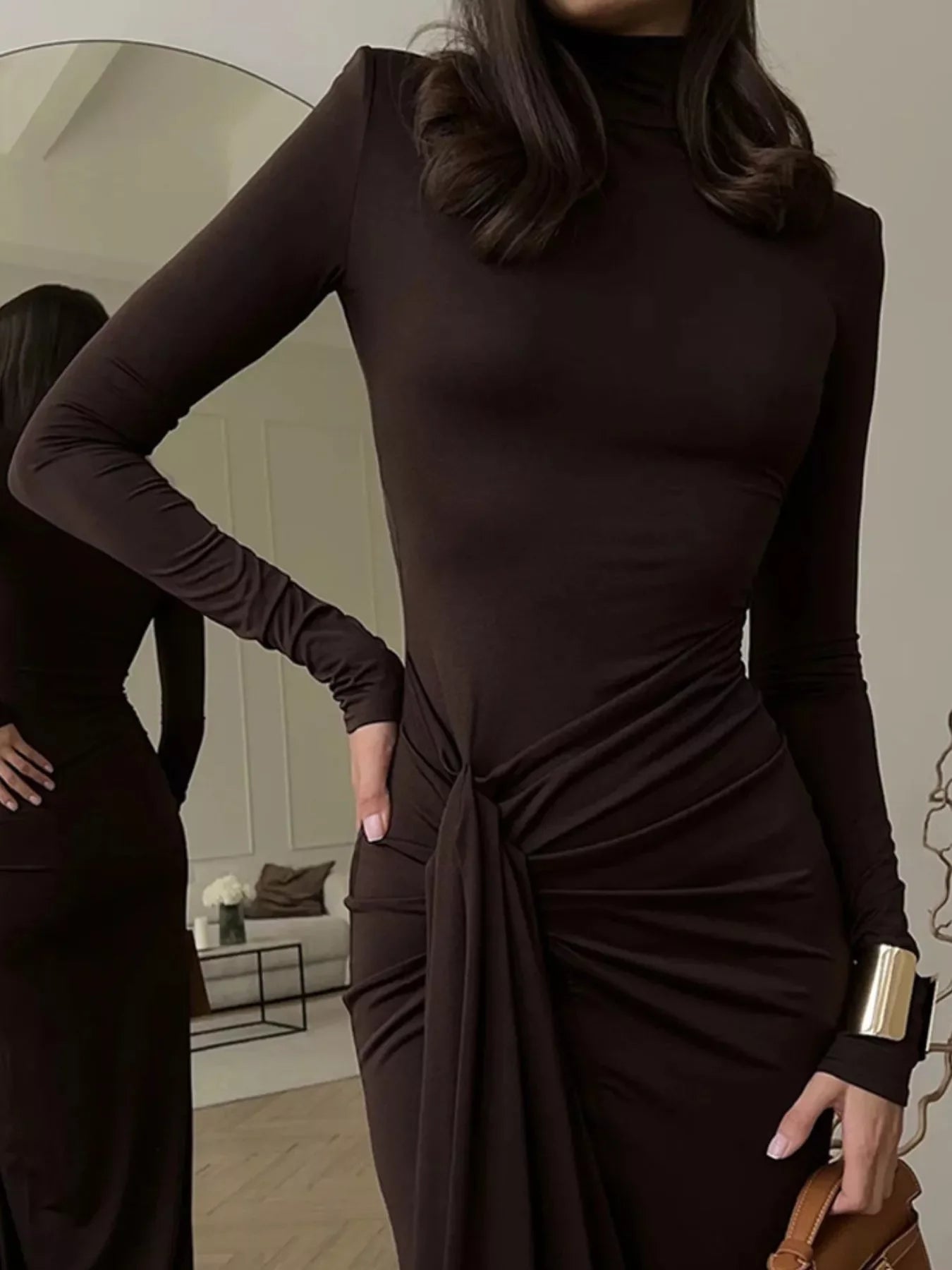 Woman wearing a fitted long-sleeve dark brown dress with draped detail, gold cuff bracelet, and holding a handbag