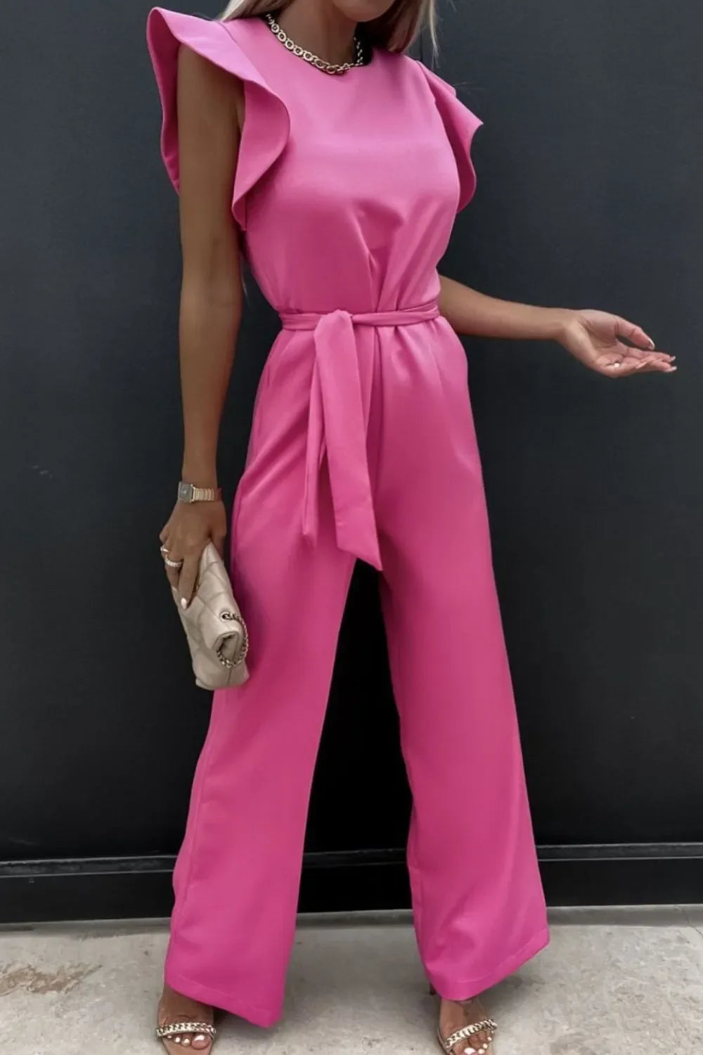 Woman wearing a pink sleeveless jumpsuit with ruffled shoulders and a waist tie, holding a beige clutch