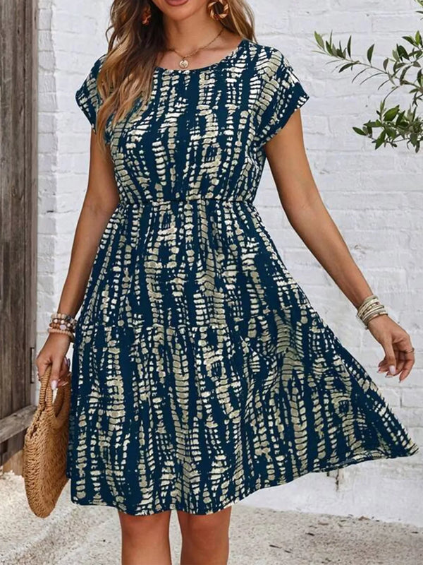 Woman wearing a blue and beige patterned short-sleeve casual dress holding a woven handbag by white brick wall