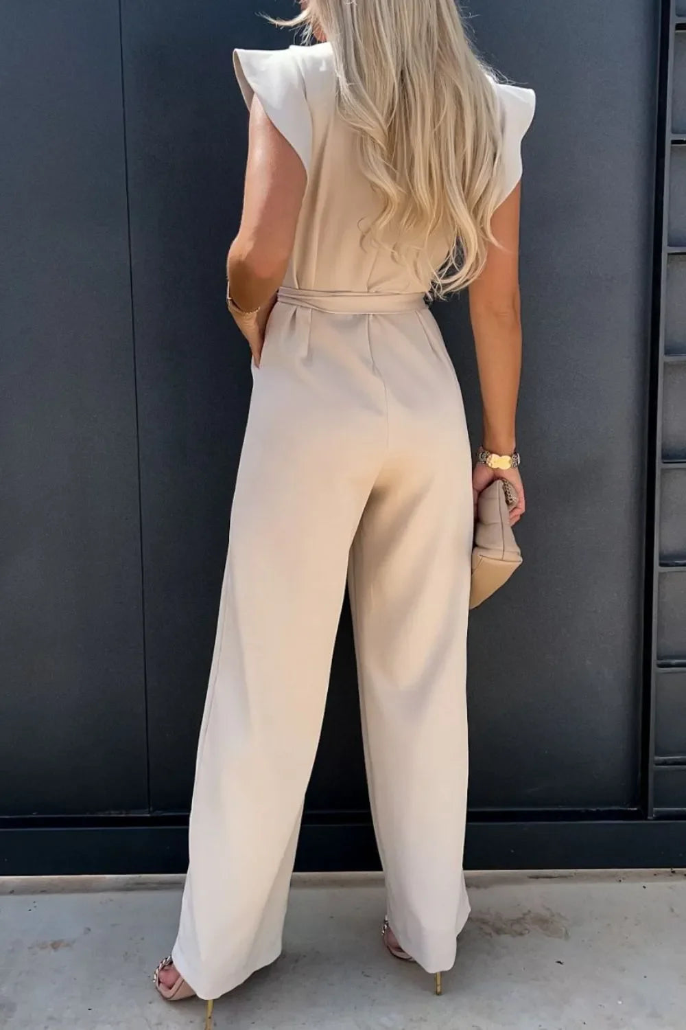 Woman in beige wide-leg pantsuit and heels holding a beige clutch, standing against dark wall