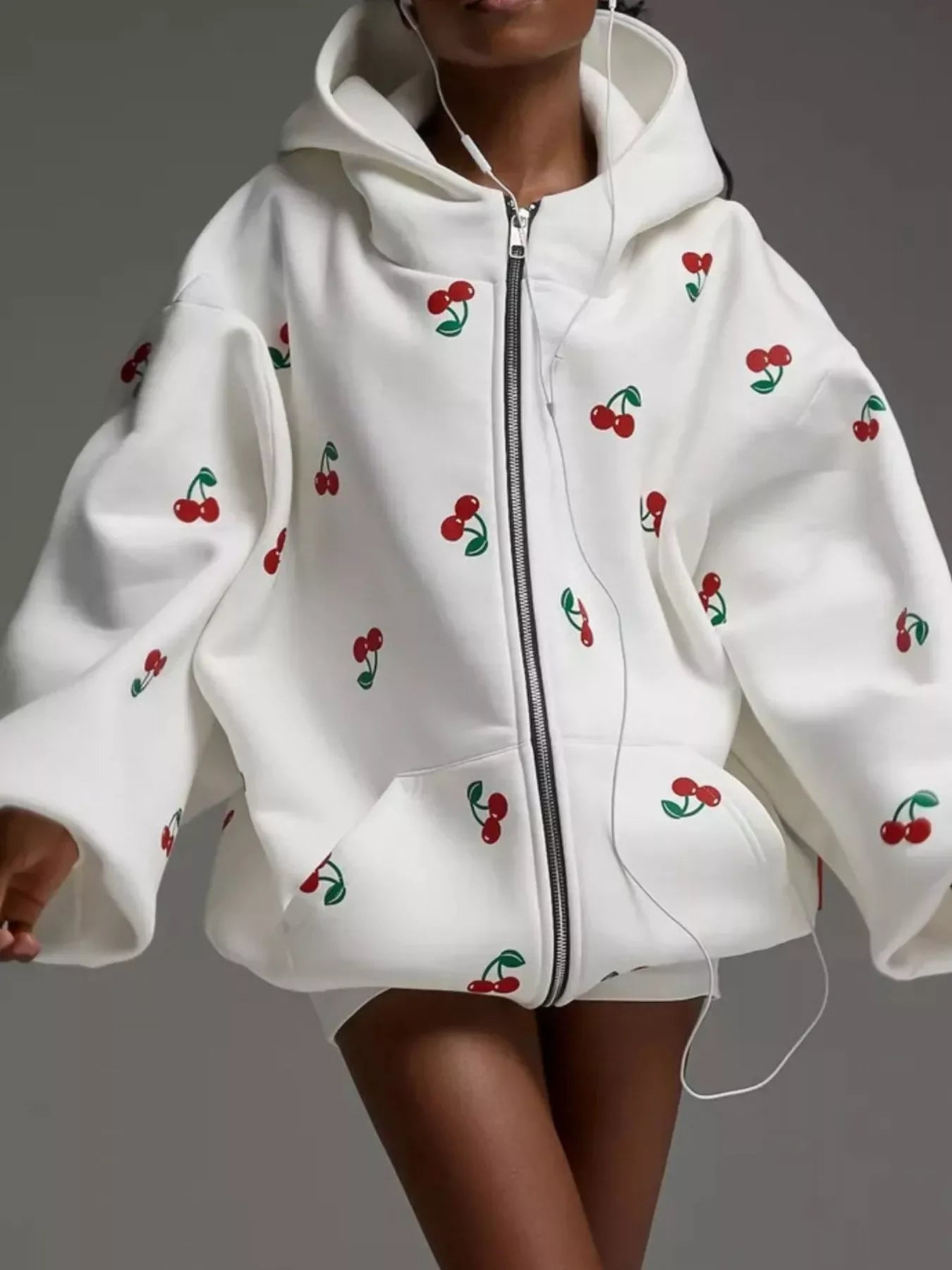White zip-up hoodie with red cherry print and oversized sleeves worn by person with earbuds