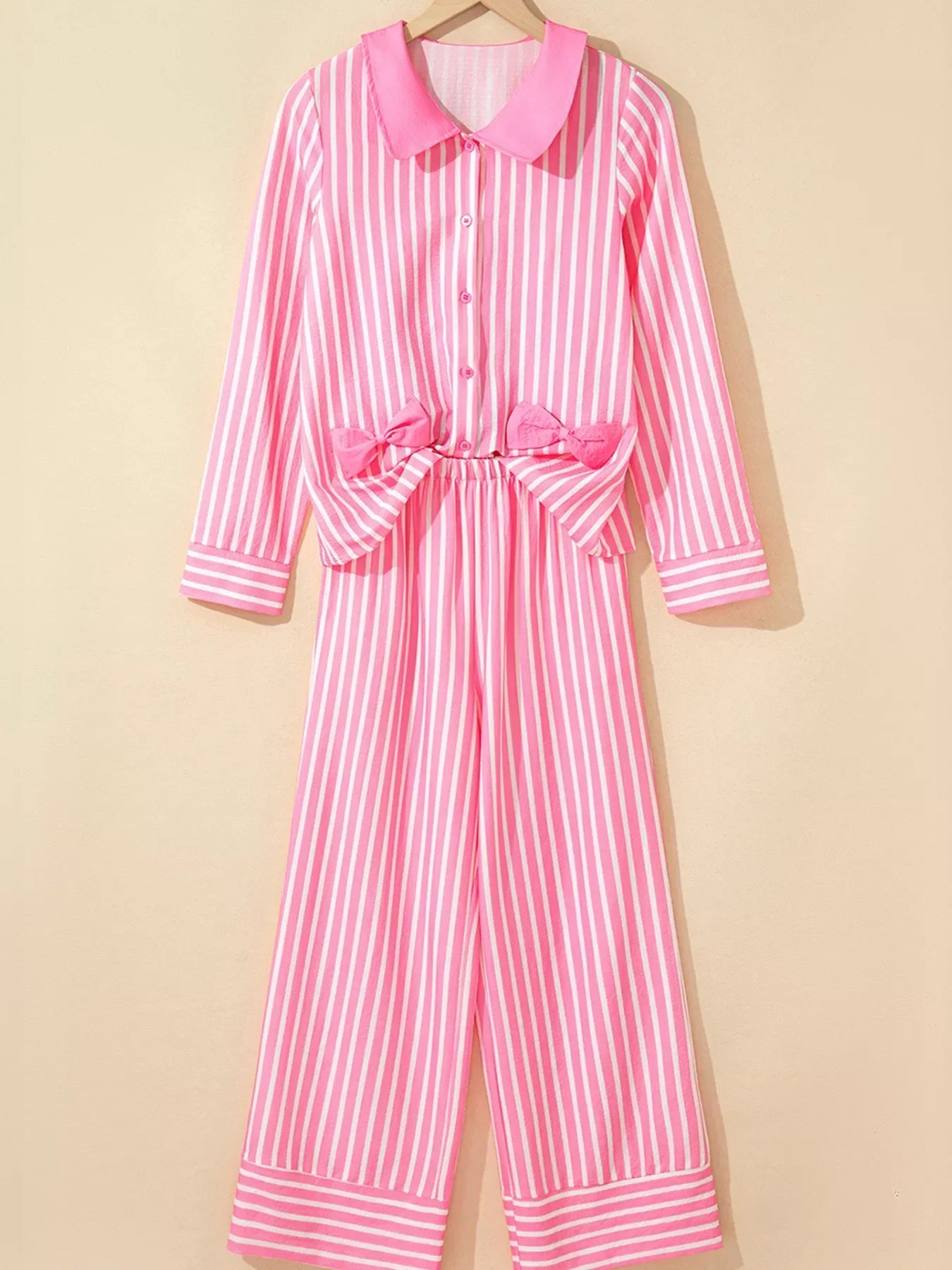 Pink and white striped long-sleeve pajama set with collar and bows on top, hanging on beige wall