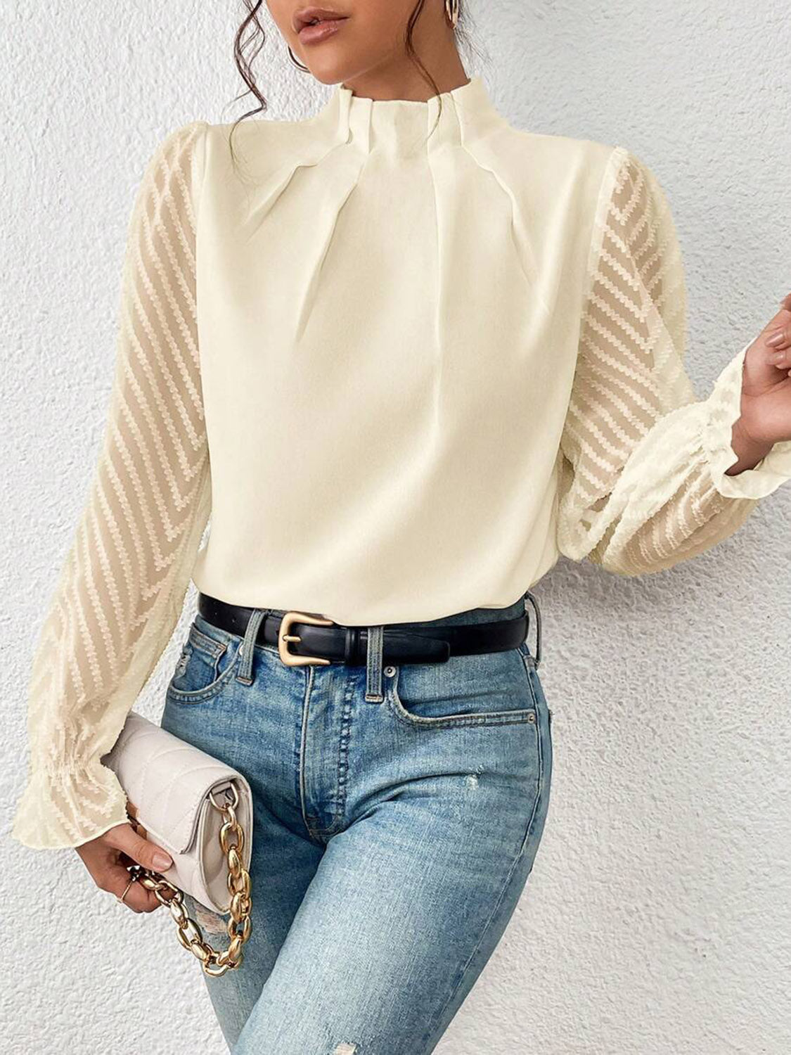 Woman wearing cream blouse with sheer patterned sleeves, blue jeans, black belt, and holding quilted handbag