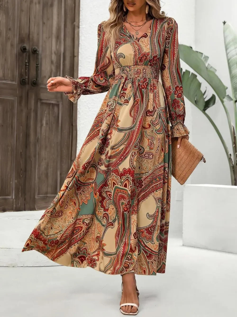 Woman wearing boho long sleeve floral paisley maxi dress with smocked waist, holding woven clutch