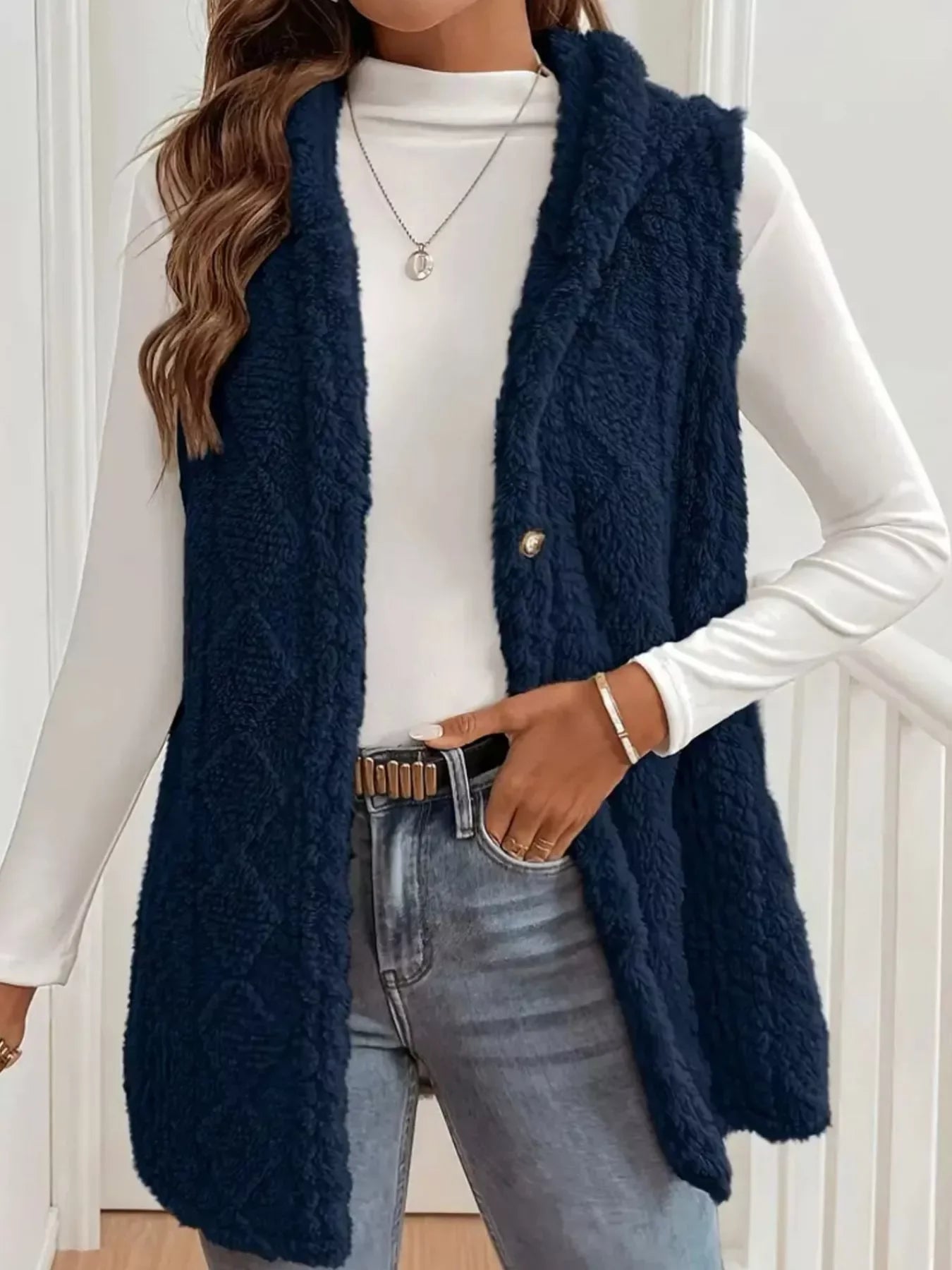 Woman wearing navy blue fuzzy vest over white long-sleeve shirt with jeans and gold accessories