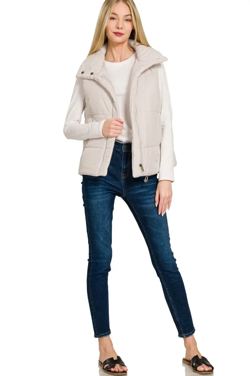 Woman wearing beige puffer vest over white long sleeve top paired with dark blue skinny jeans and black slide sandals
