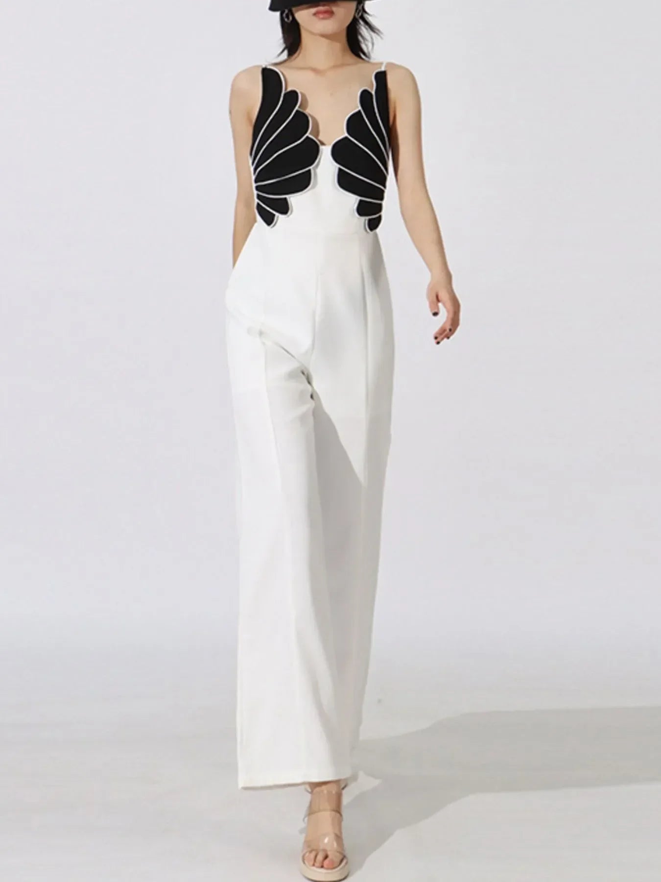 Woman modeling white wide-leg jumpsuit with black scallop design on bodice and clear heels