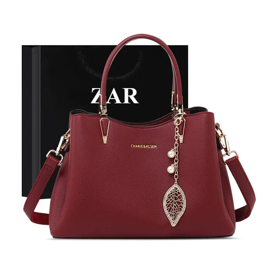 Burgundy leather handbag with gold hardware and leaf charm, against a black gift bag
