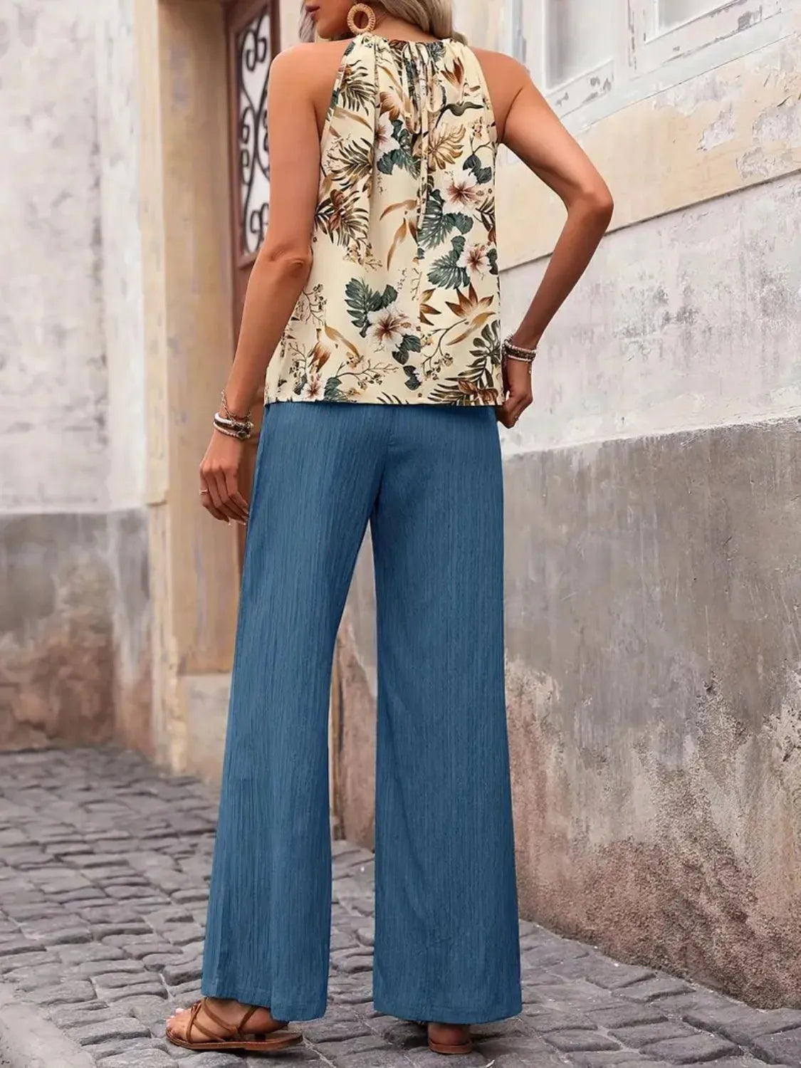 Woman wearing floral sleeveless top and loose blue pants, standing outdoors on cobblestone street