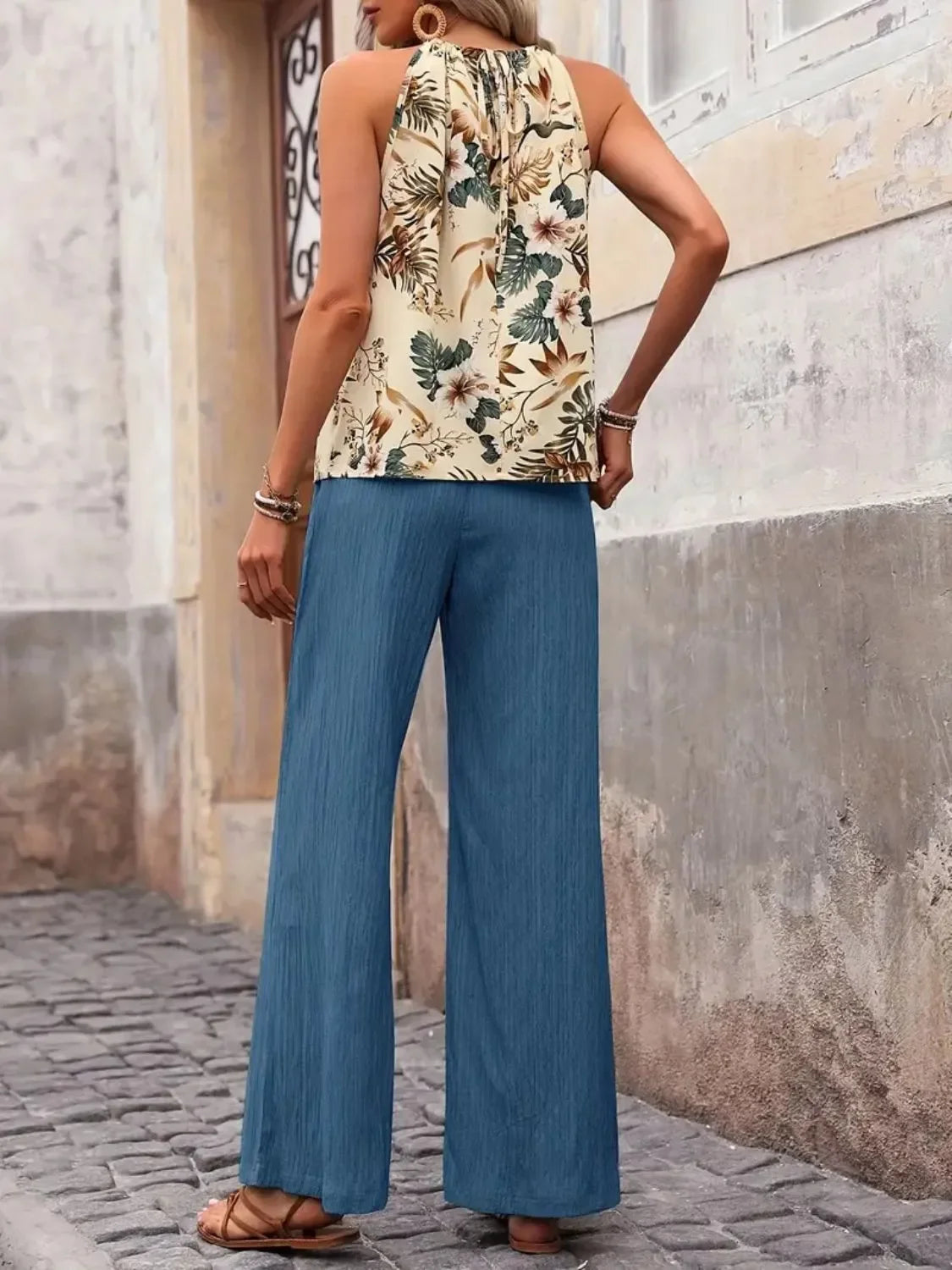 Woman wearing floral sleeveless top and loose blue pants, standing outdoors on cobblestone street