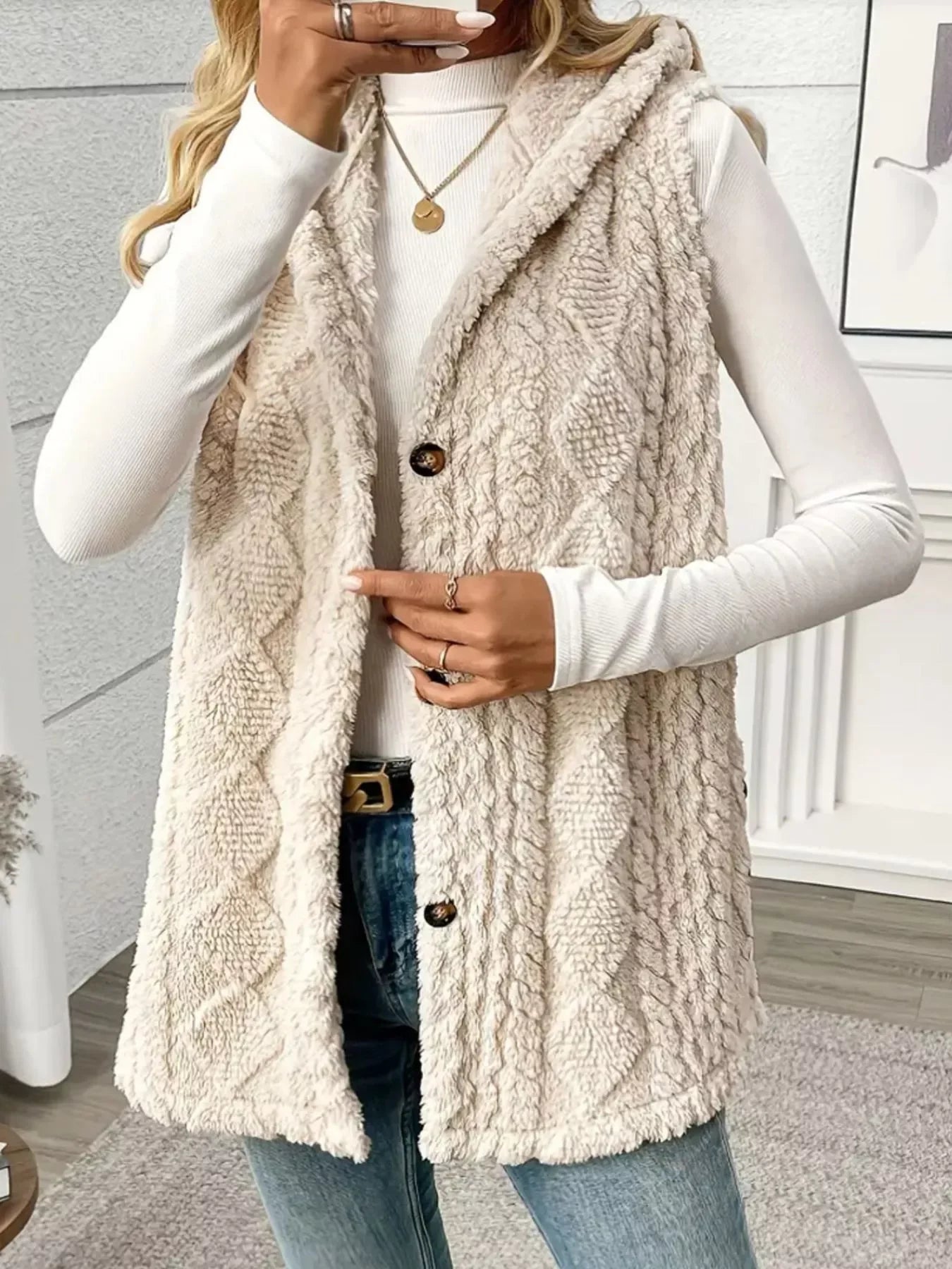 Woman wearing a beige textured faux fur vest over a white long sleeve top with jeans indoors