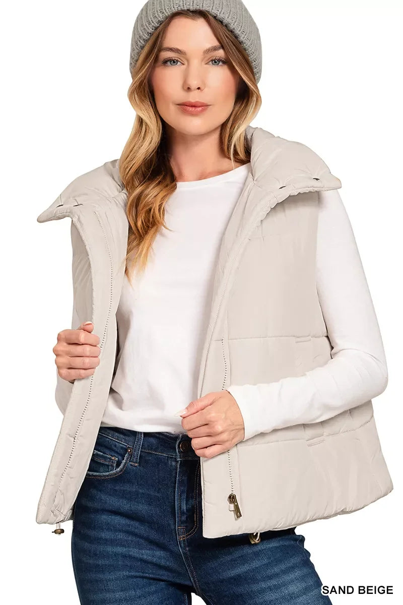 Woman wearing sand beige puffer vest, white long-sleeve top, gray beanie, and blue jeans