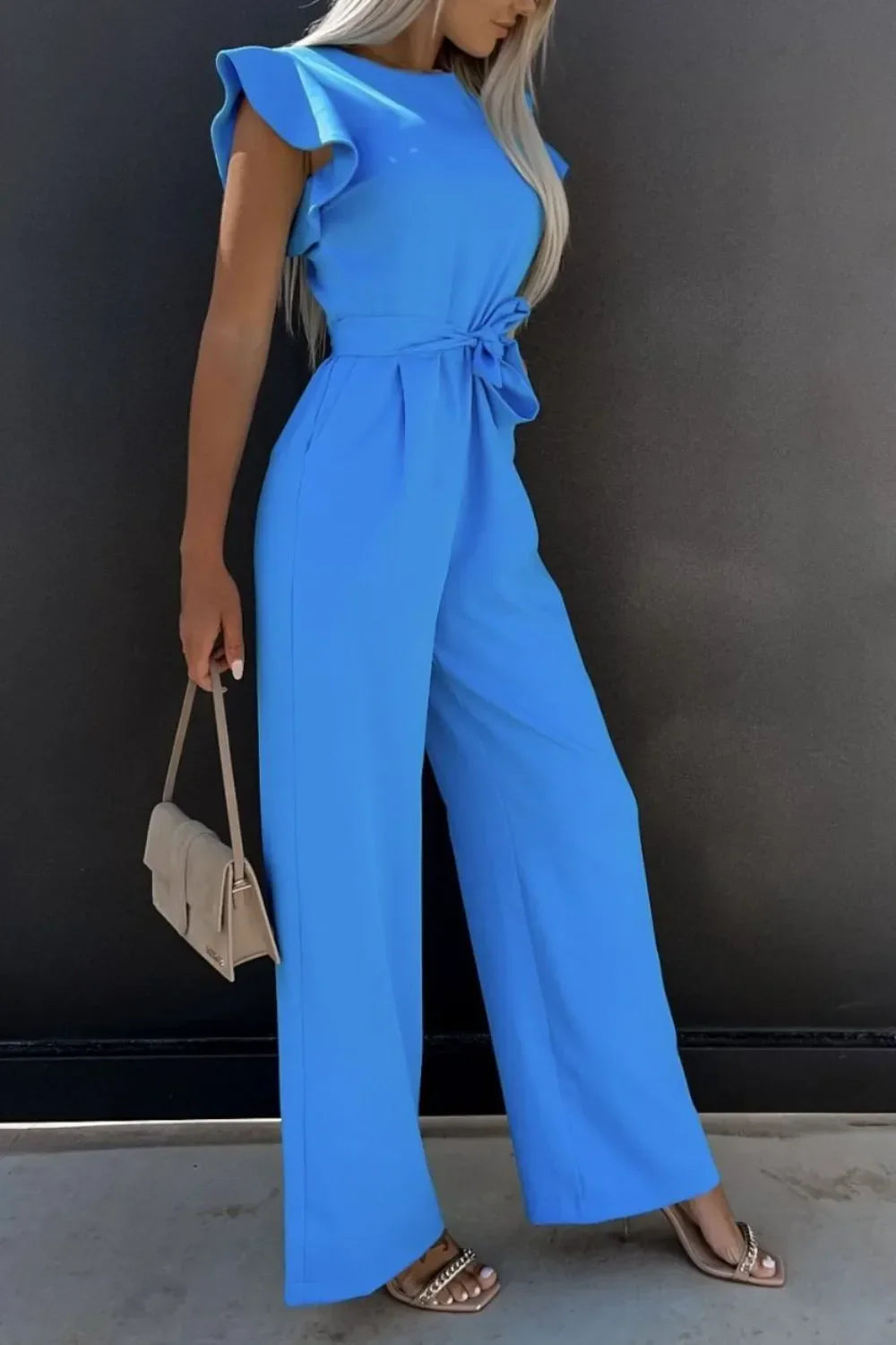 Woman in a bright blue ruffle sleeve jumpsuit with wide legs holding a beige handbag and wearing chain strap sandals