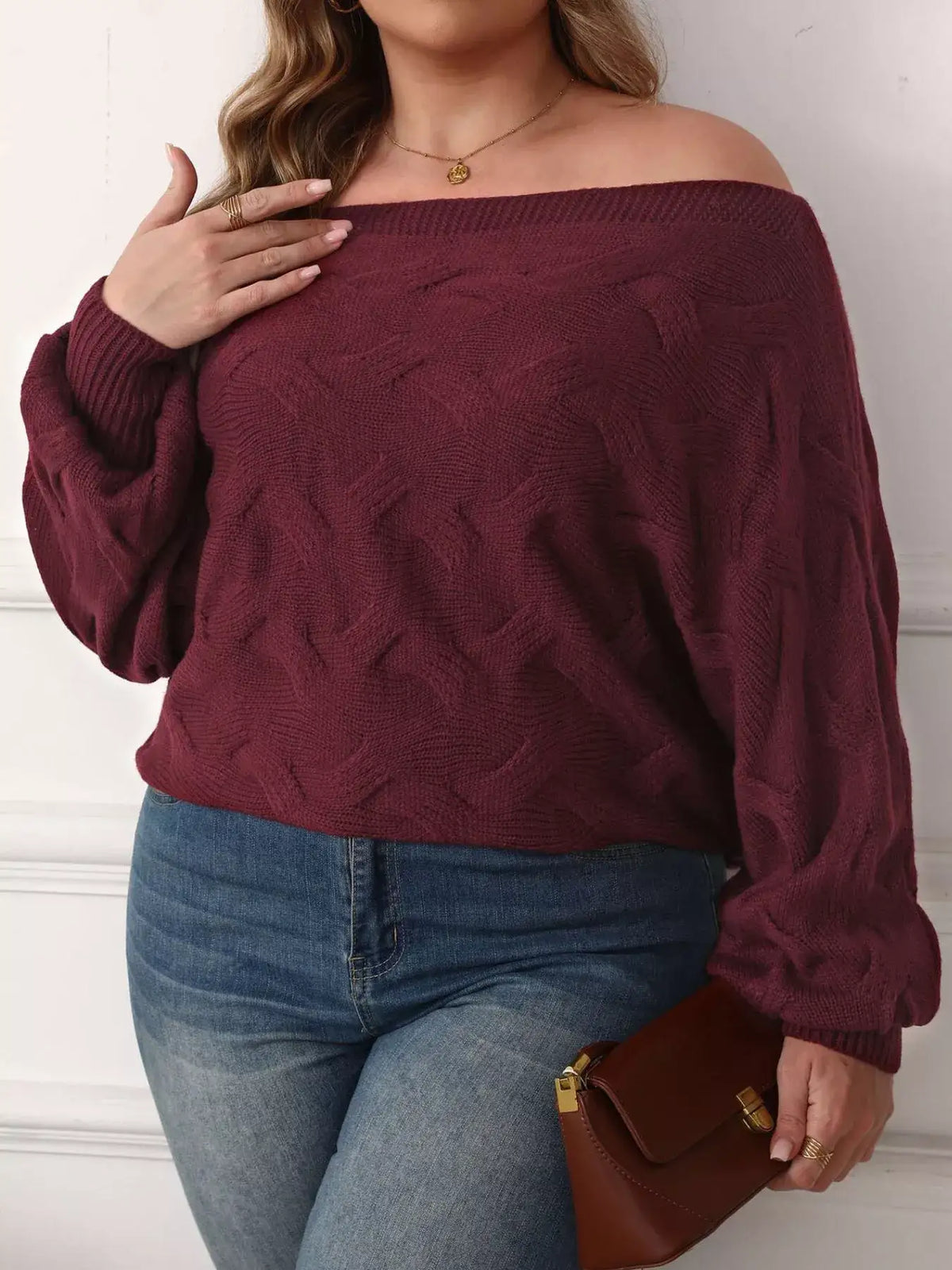 Plus size burgundy off-shoulder cable knit sweater with long sleeves paired with blue jeans.