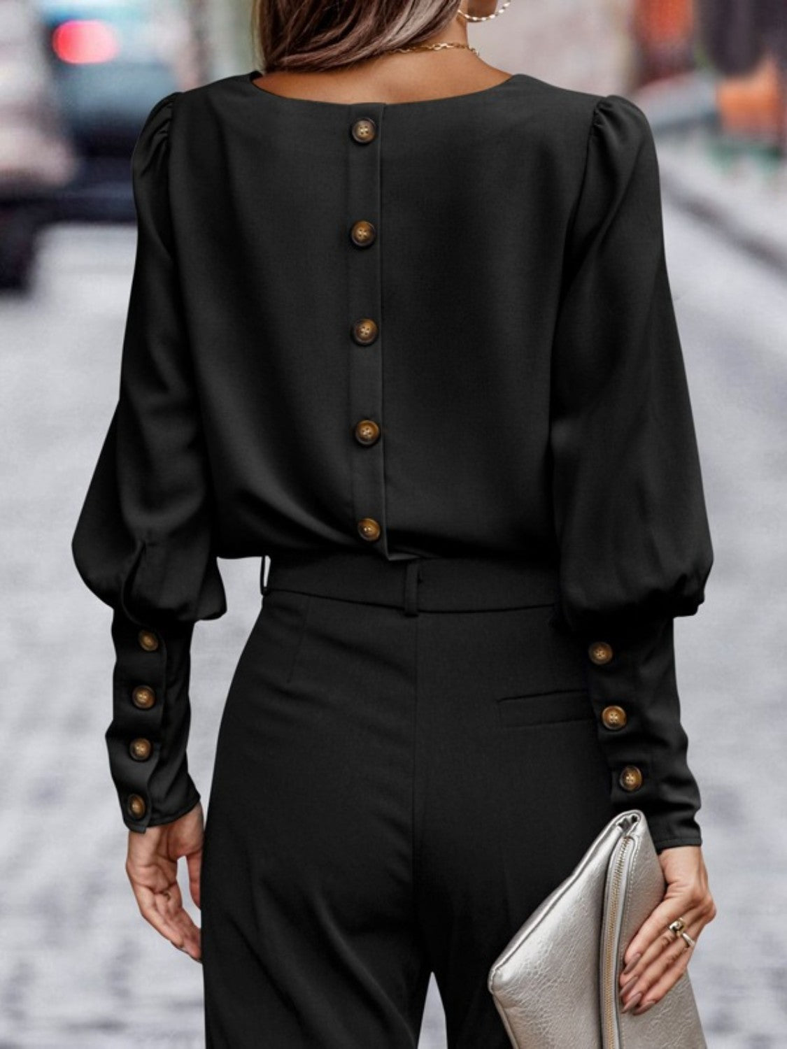 Woman in black button-back blouse and high-waist pants holding metallic clutch on city street