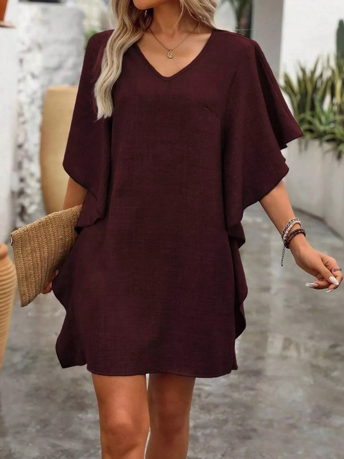 Woman wearing a loose burgundy V-neck casual dress, holding a woven clutch outdoors.