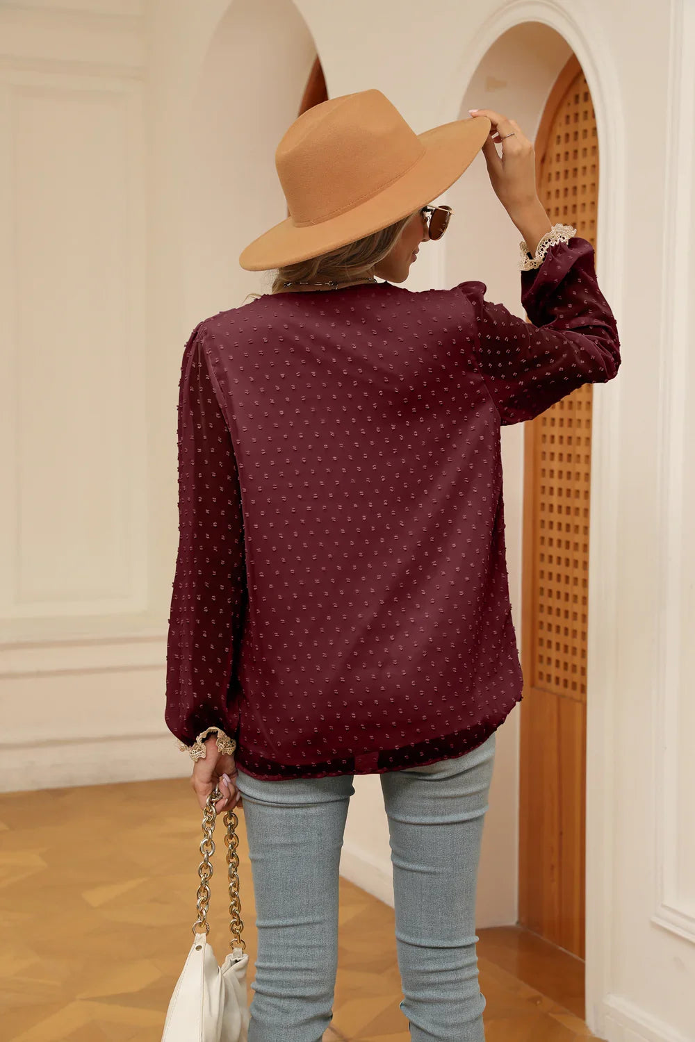 Woman in a burgundy textured blouse, wide-brim tan hat, light jeans, holding white handbag indoors