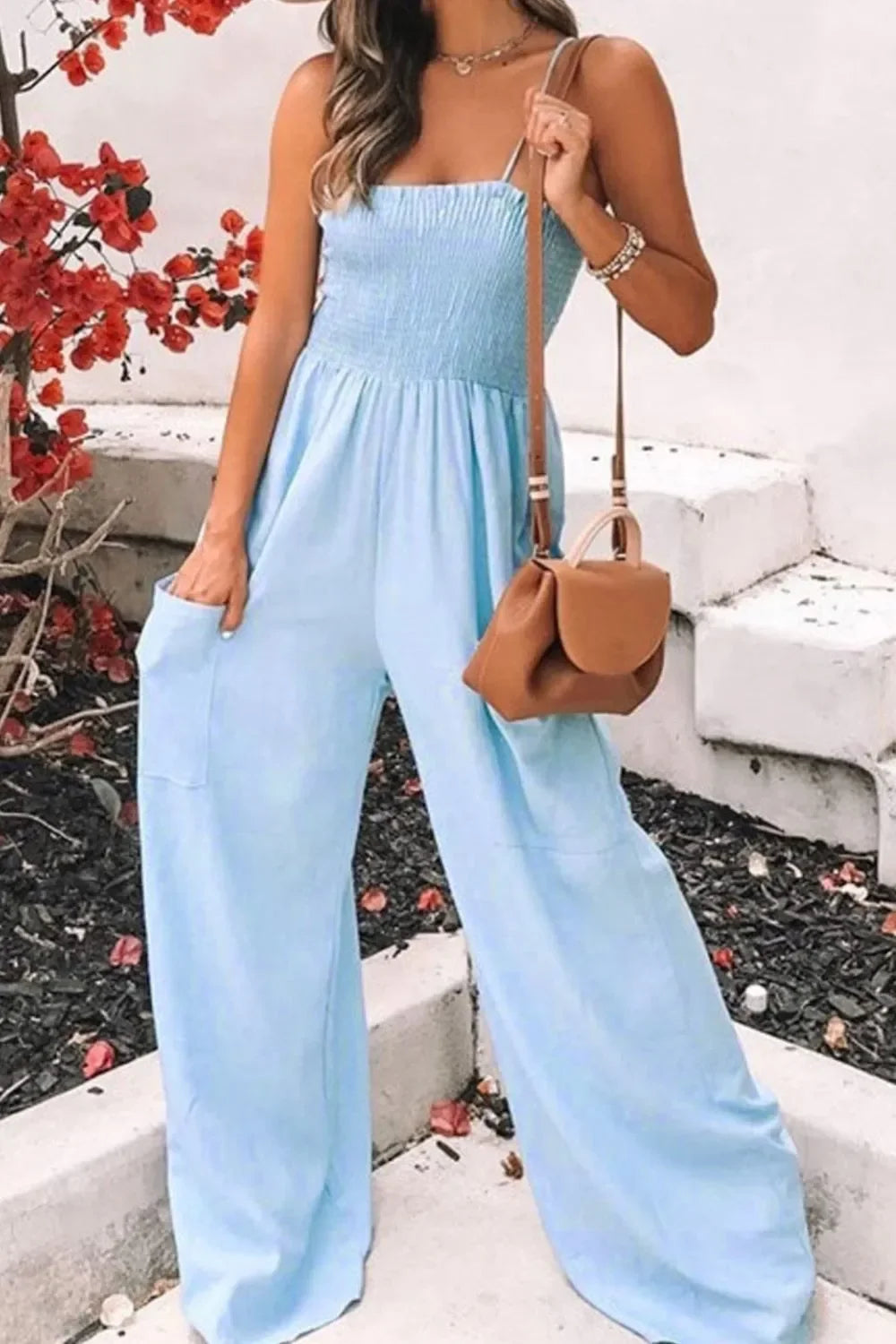 Woman wearing light blue wide-leg jumpsuit with smocked top and carrying brown handbag outdoors