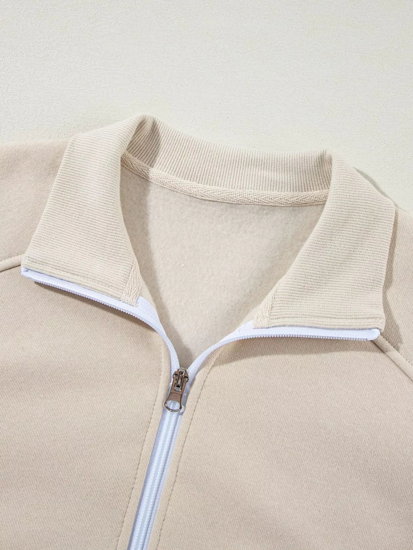 Beige fleece jacket with ribbed collar and white zipper detail, close-up view