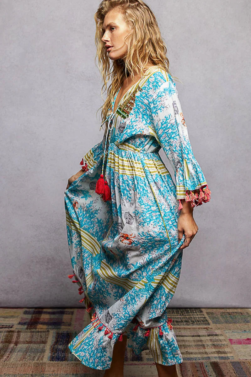 Woman in a flowy blue and yellow floral maxi dress with tassel details and flared sleeves.