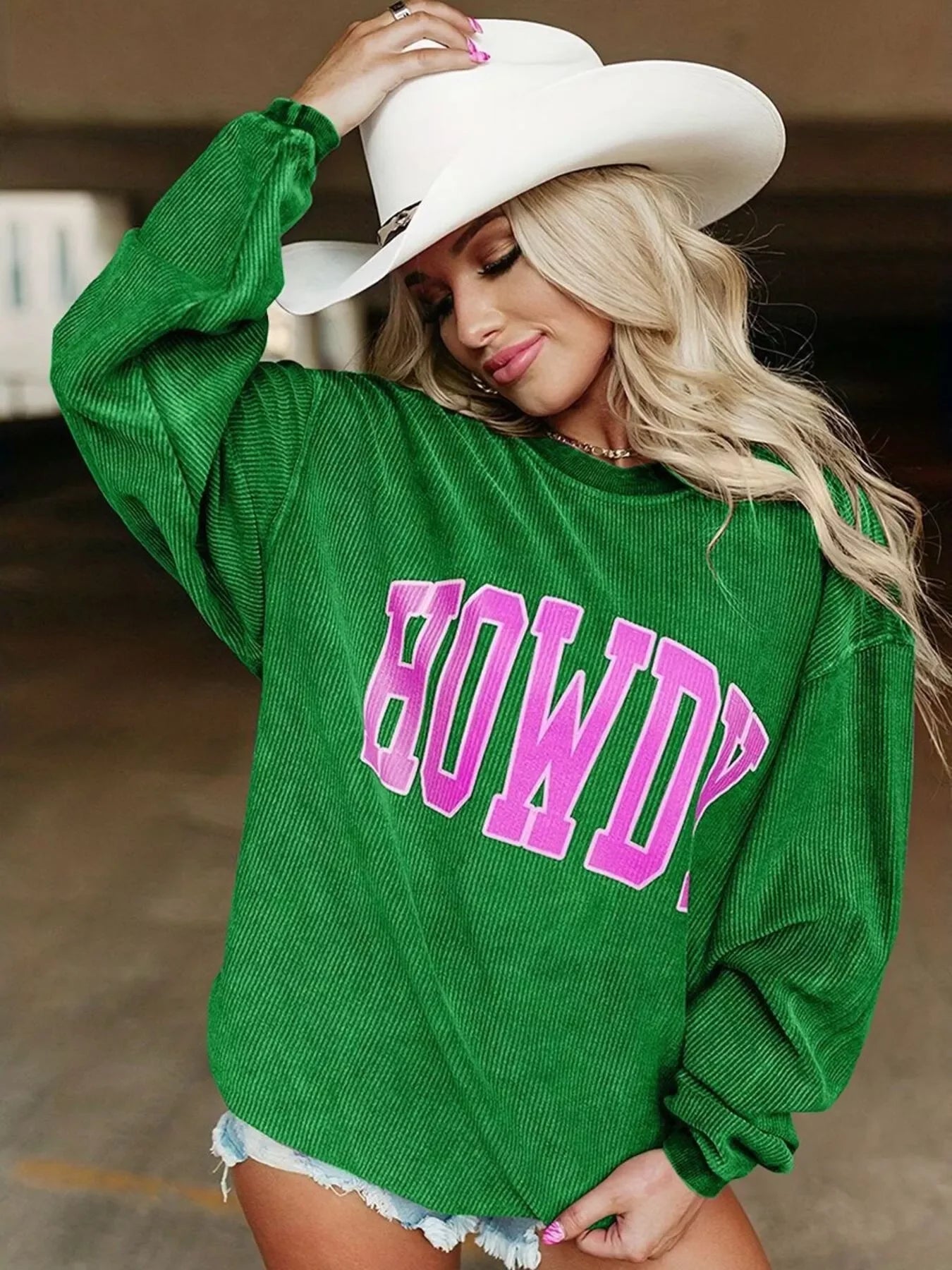 Woman wearing green corduroy sweatshirt with pink HOWDY text and white cowboy hat