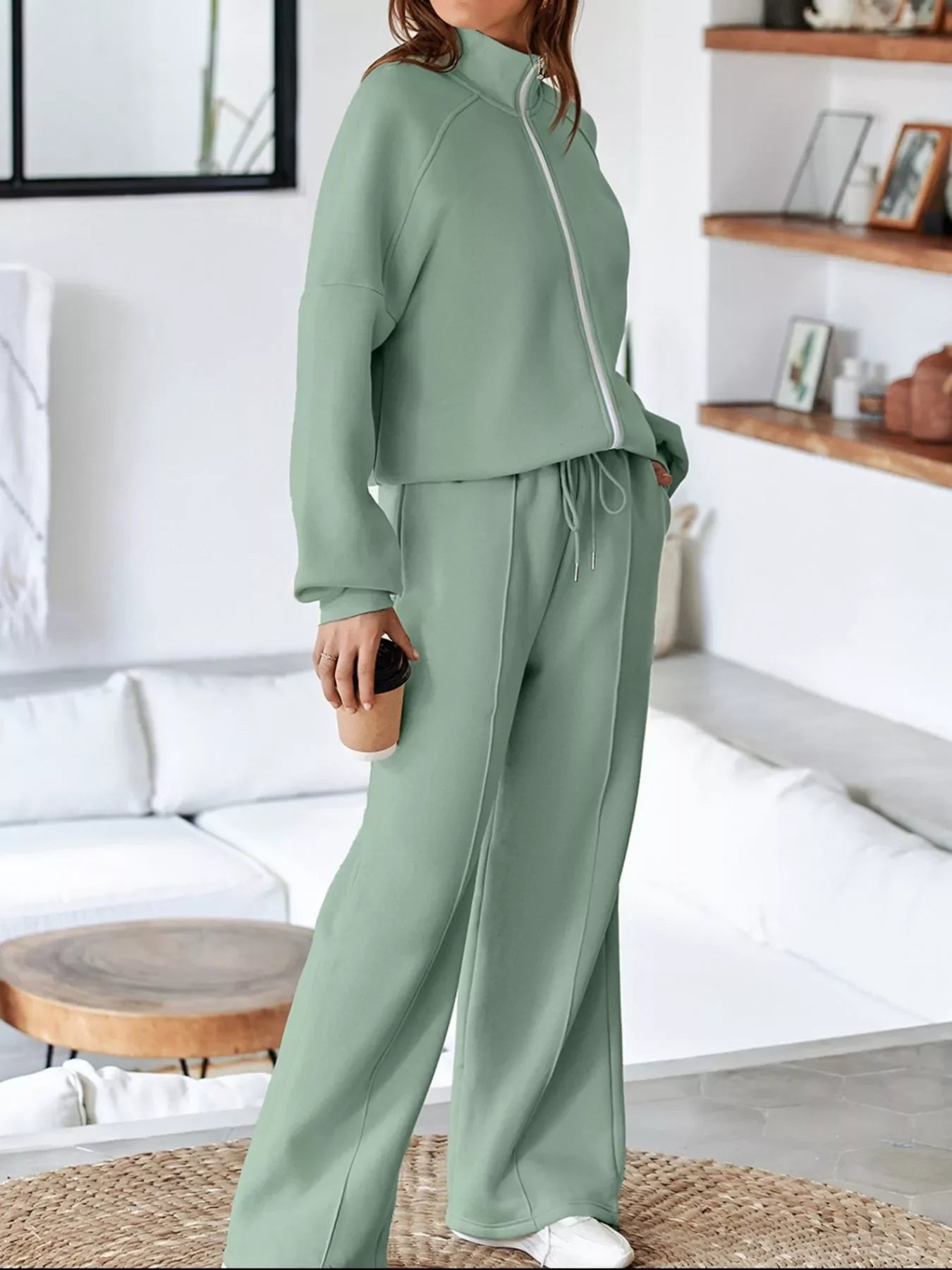 Woman in sage green zip-up sweatshirt and matching wide-leg pants holding coffee cup in cozy modern living room
