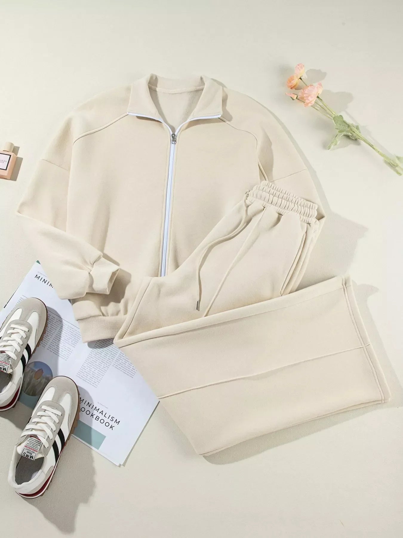 Beige zip-up sweatshirt and matching sweatpants laid flat with sneakers and a flower on a light background