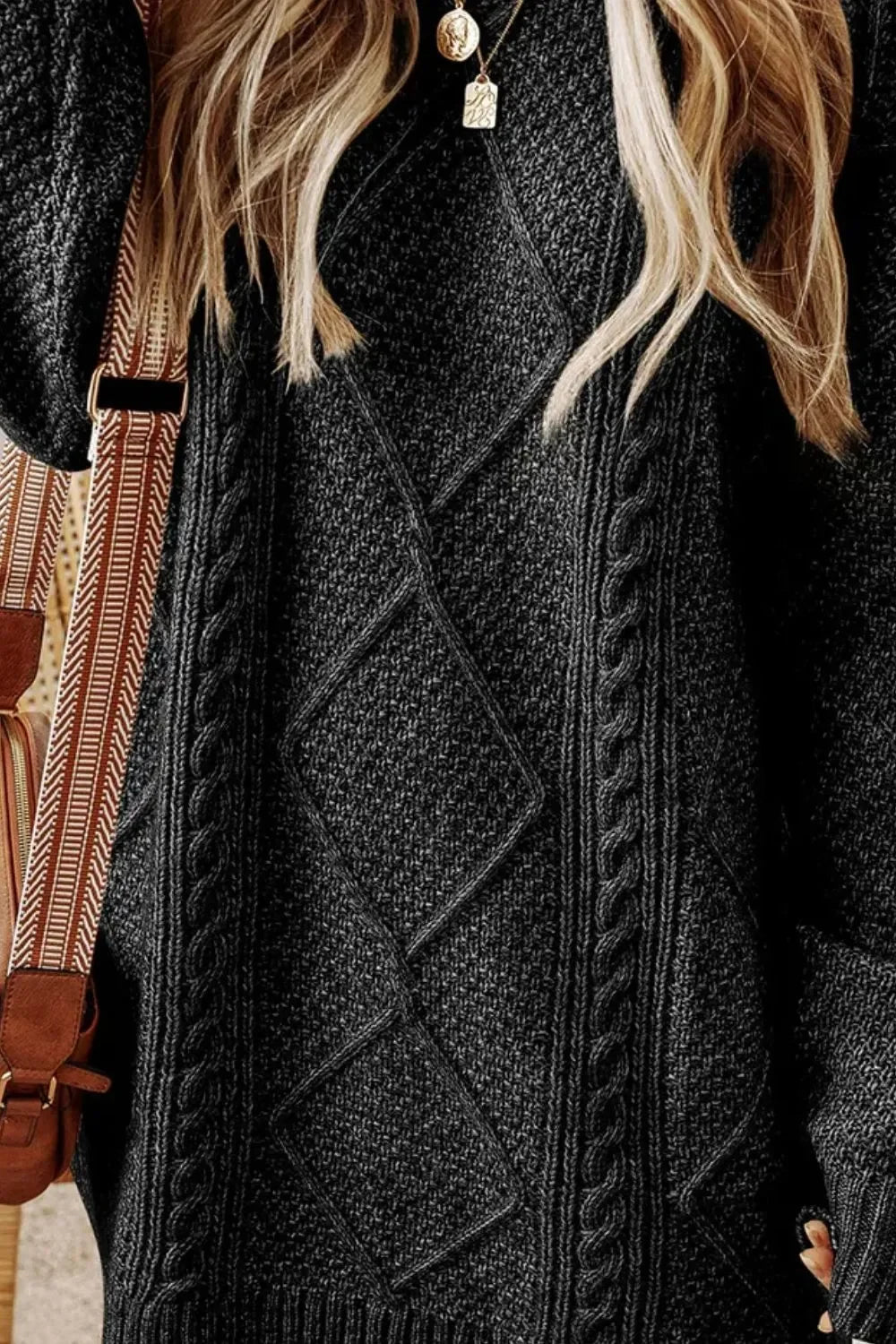 Close-up of a woman wearing a textured dark gray cable knit sweater with diamond patterns, layered necklaces, and a brown strap bag
