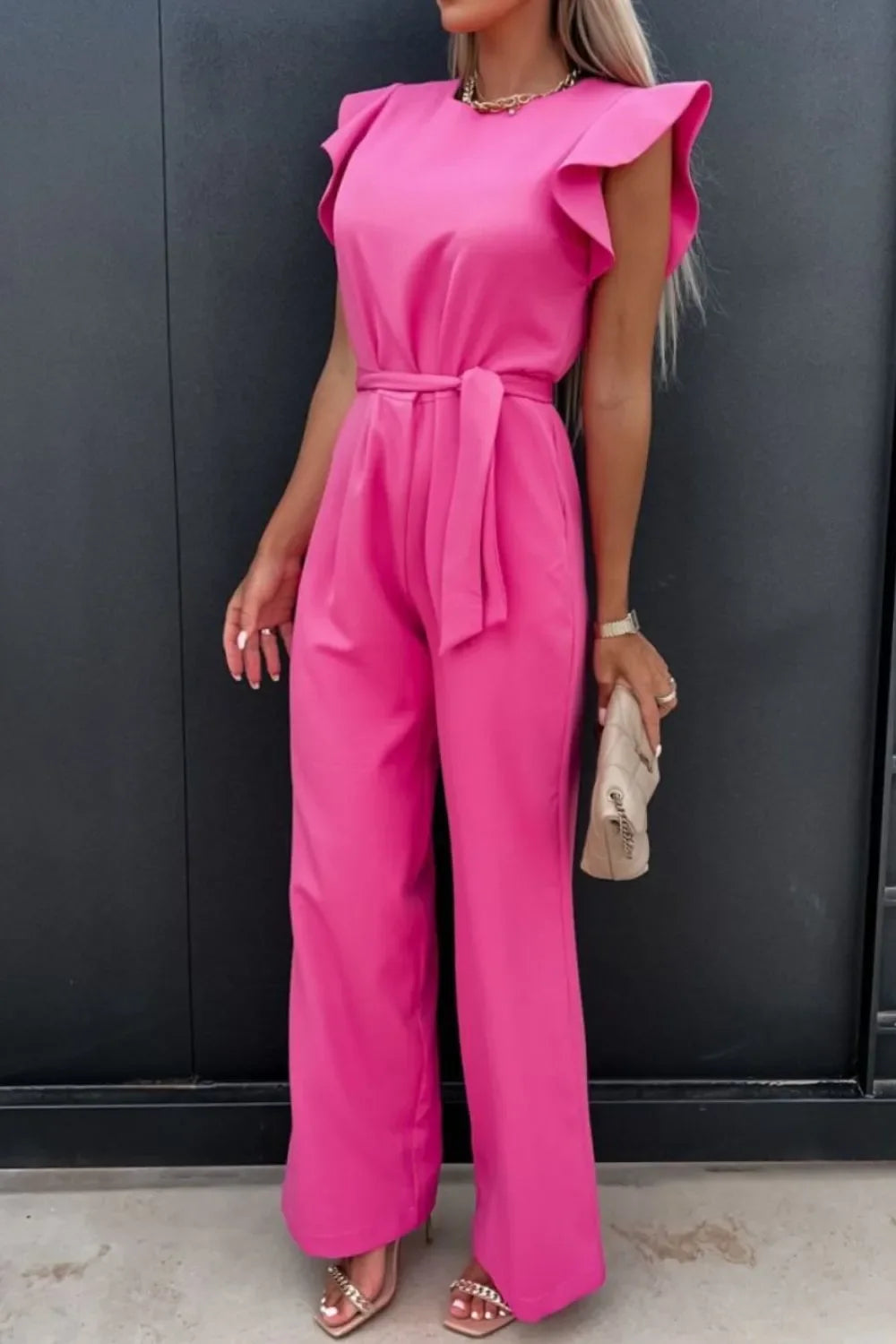 Woman in vibrant pink jumpsuit with ruffled sleeves, tied waist belt, holding beige clutch bag