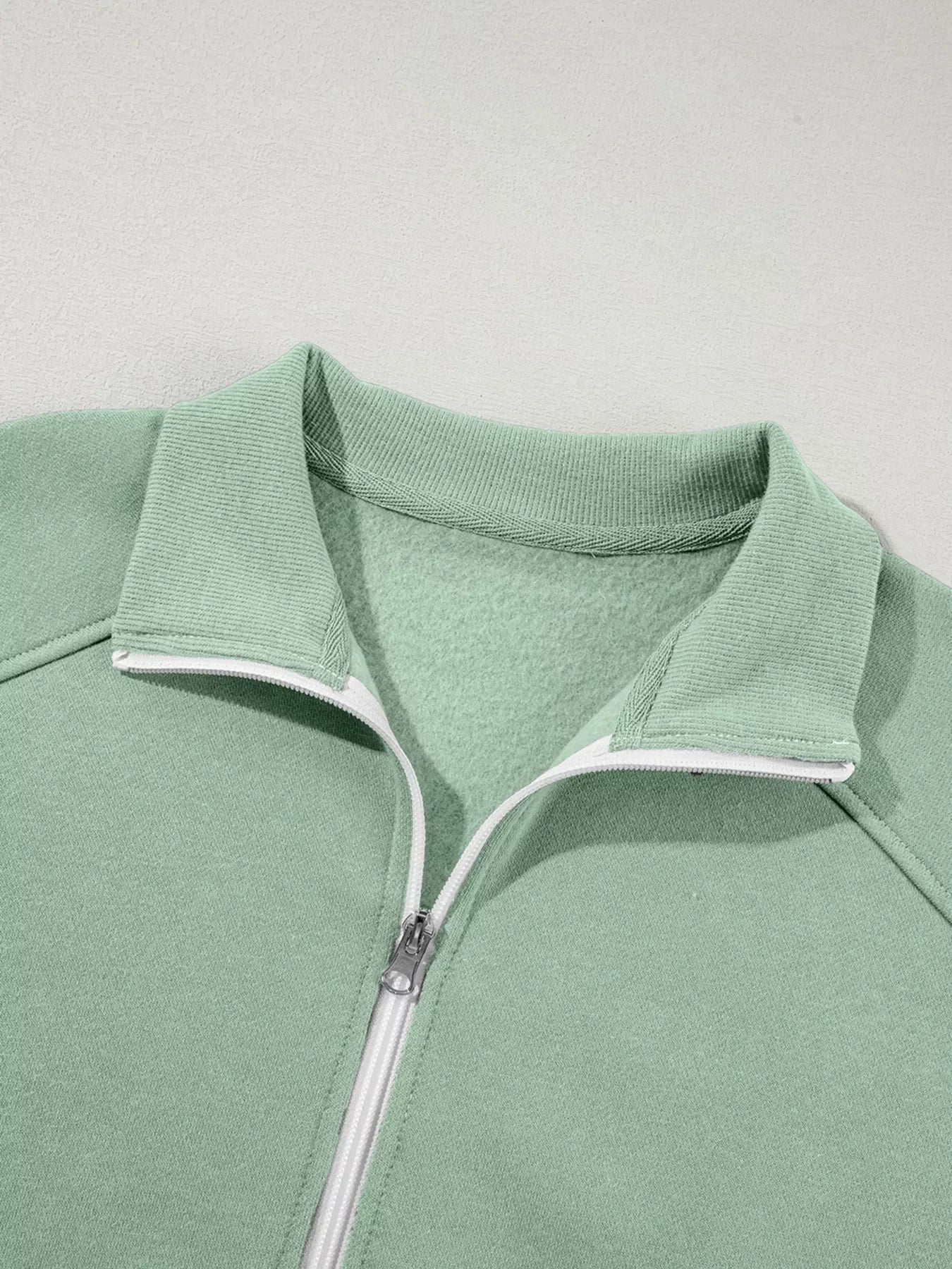 Close-up of sage green fleece jacket with white zipper and ribbed collar against white background