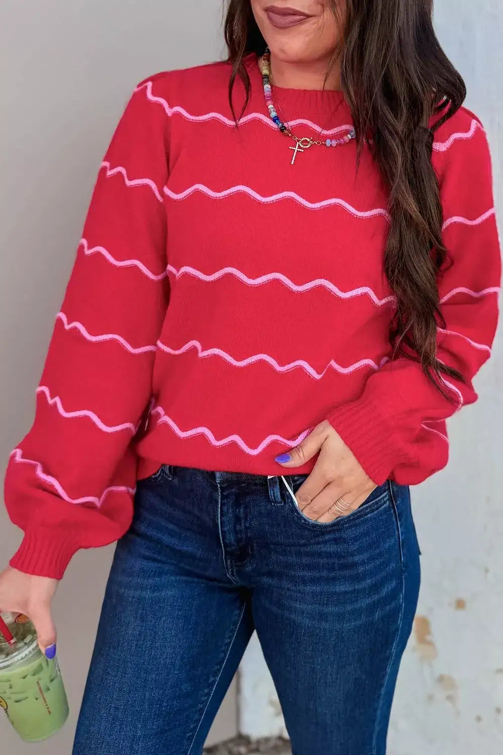 Woman wearing a red Evie's Dreamland round neck long sleeve sweater with pink wavy stripes and blue jeans.