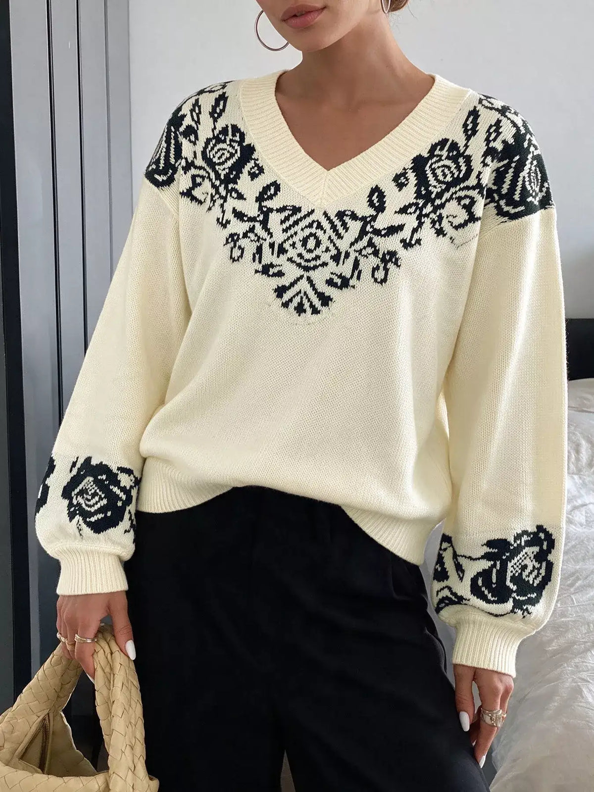 Cream V-neck dropped shoulder sweater with black floral patterns paired with black trousers.