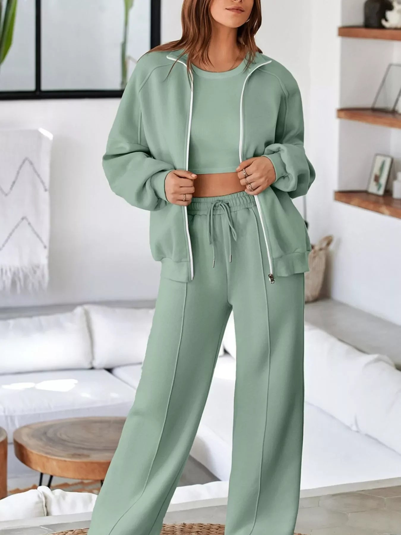 Woman wearing sage green three-piece loungewear set with zip-up jacket, crop top, and drawstring pants in modern living room