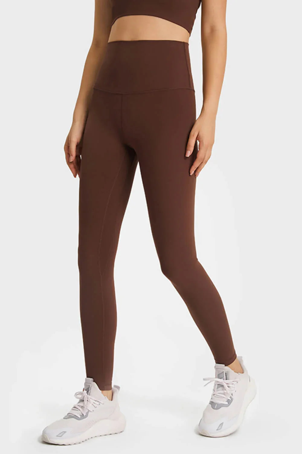 Woman wearing high-waisted brown leggings and white athletic sneakers on white background
