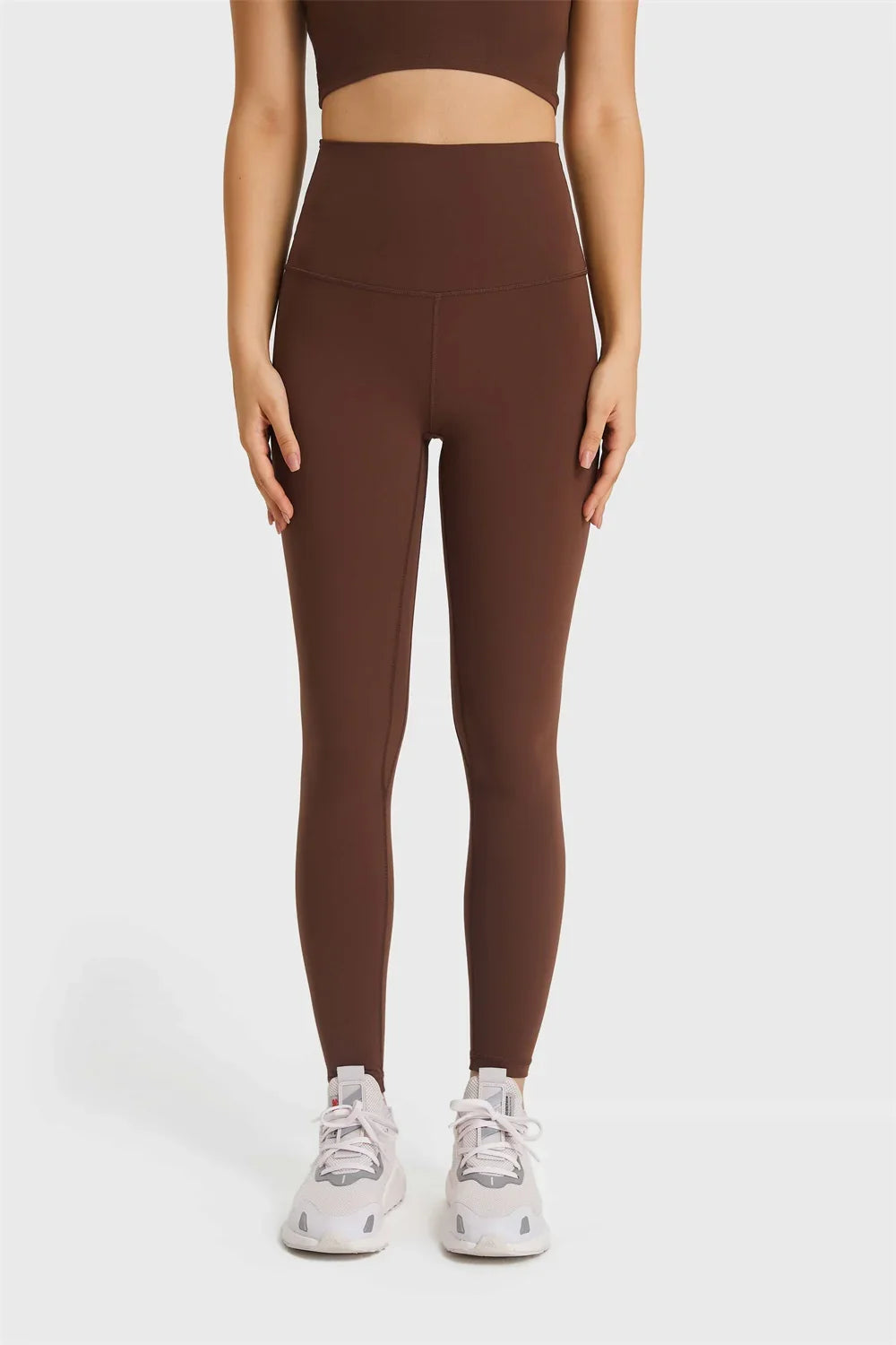 Woman wearing high-waisted brown leggings and matching crop top with white sneakers on gray background