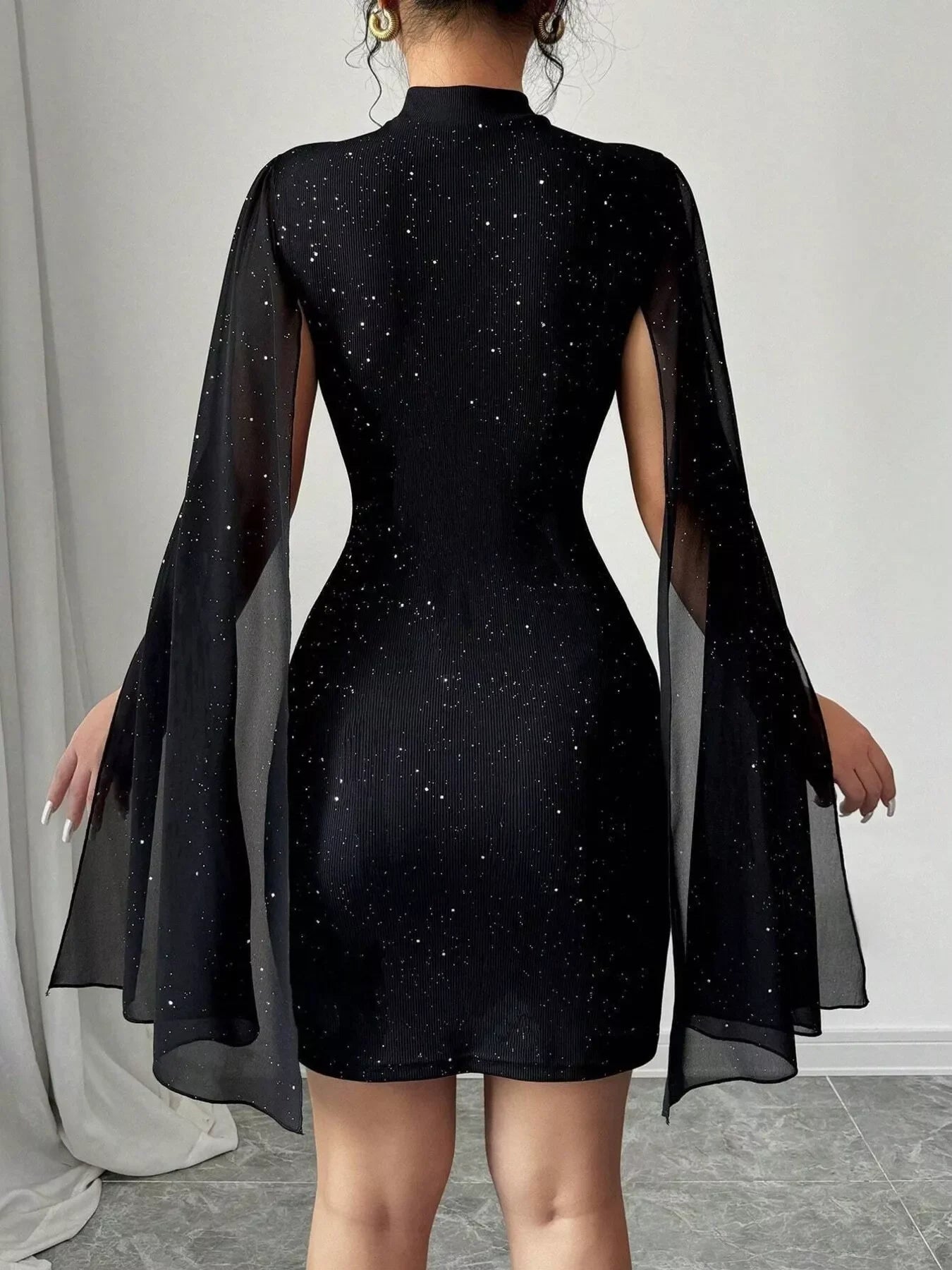 Back view of woman wearing a black glitter mini dress with flowy long sheer sleeves and gold hoop earrings