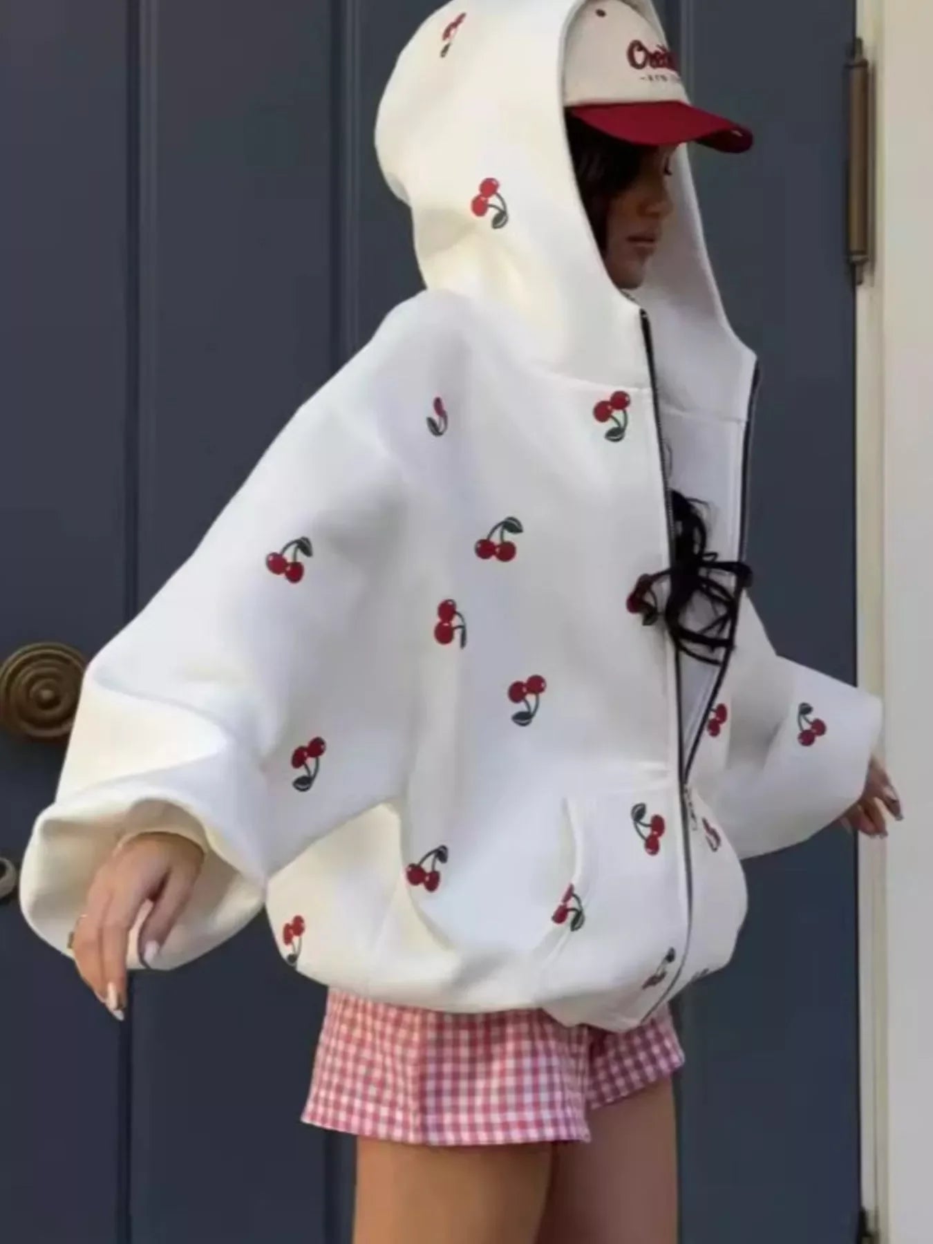 Woman wearing oversized white hoodie with red cherry print and red gingham shorts, standing by a door