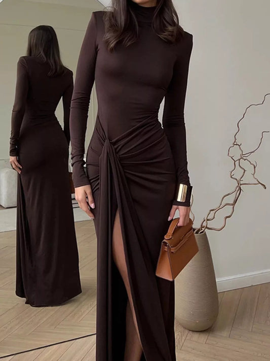 Woman in long-sleeve dark brown maxi dress with high slit and brown handbag in modern room